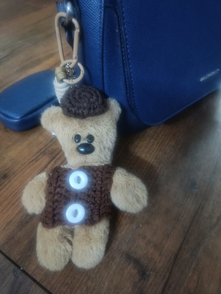 Blue sling bag with a small teddy bear keychain resting on a wooden surface.
